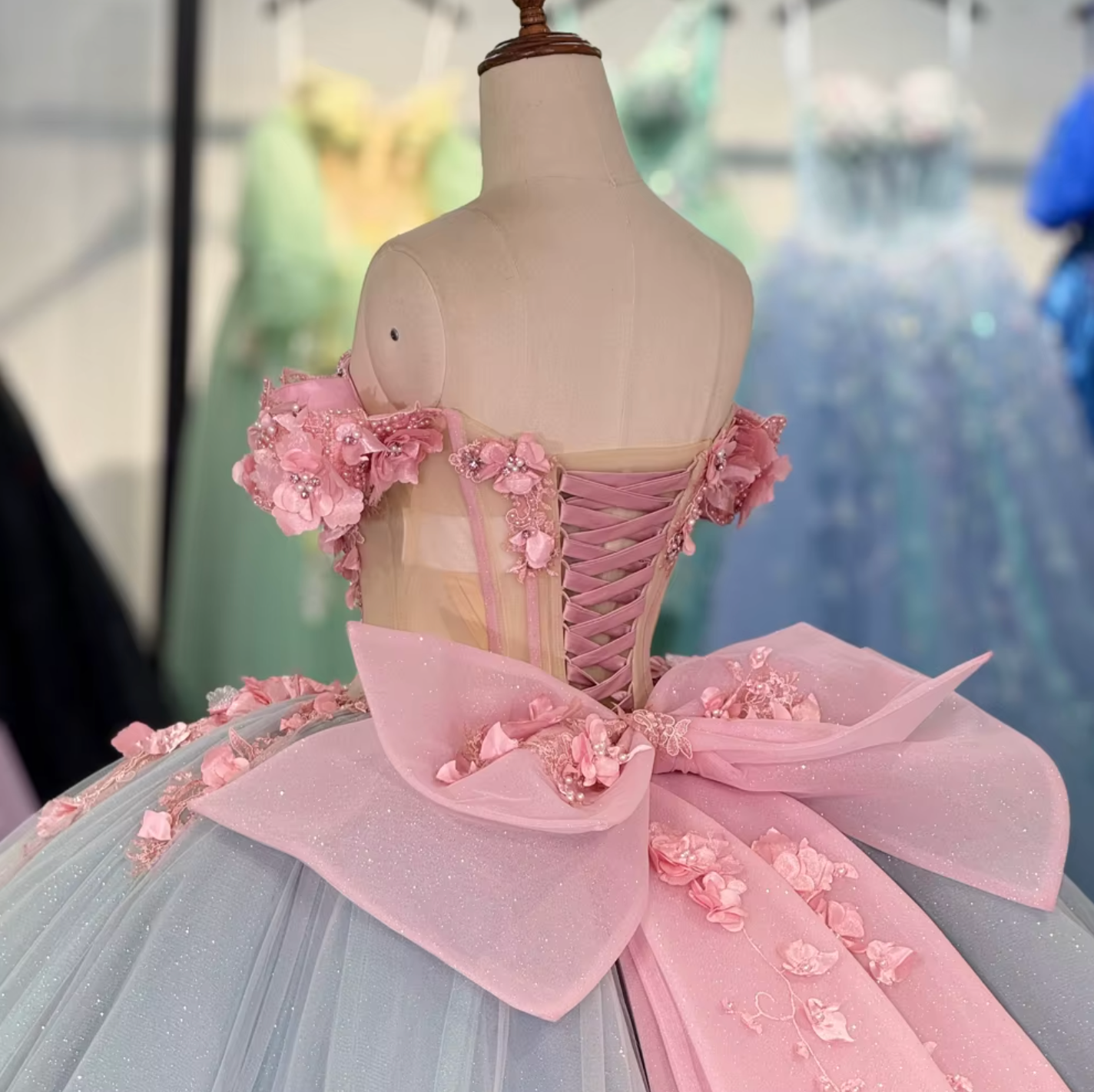 A stunning blue and pink Quinceanera dress with a sheer lace bodice, 3D floral decorations, a sweetheart neckline, and a full ballroom skirt with an oversized back bow.