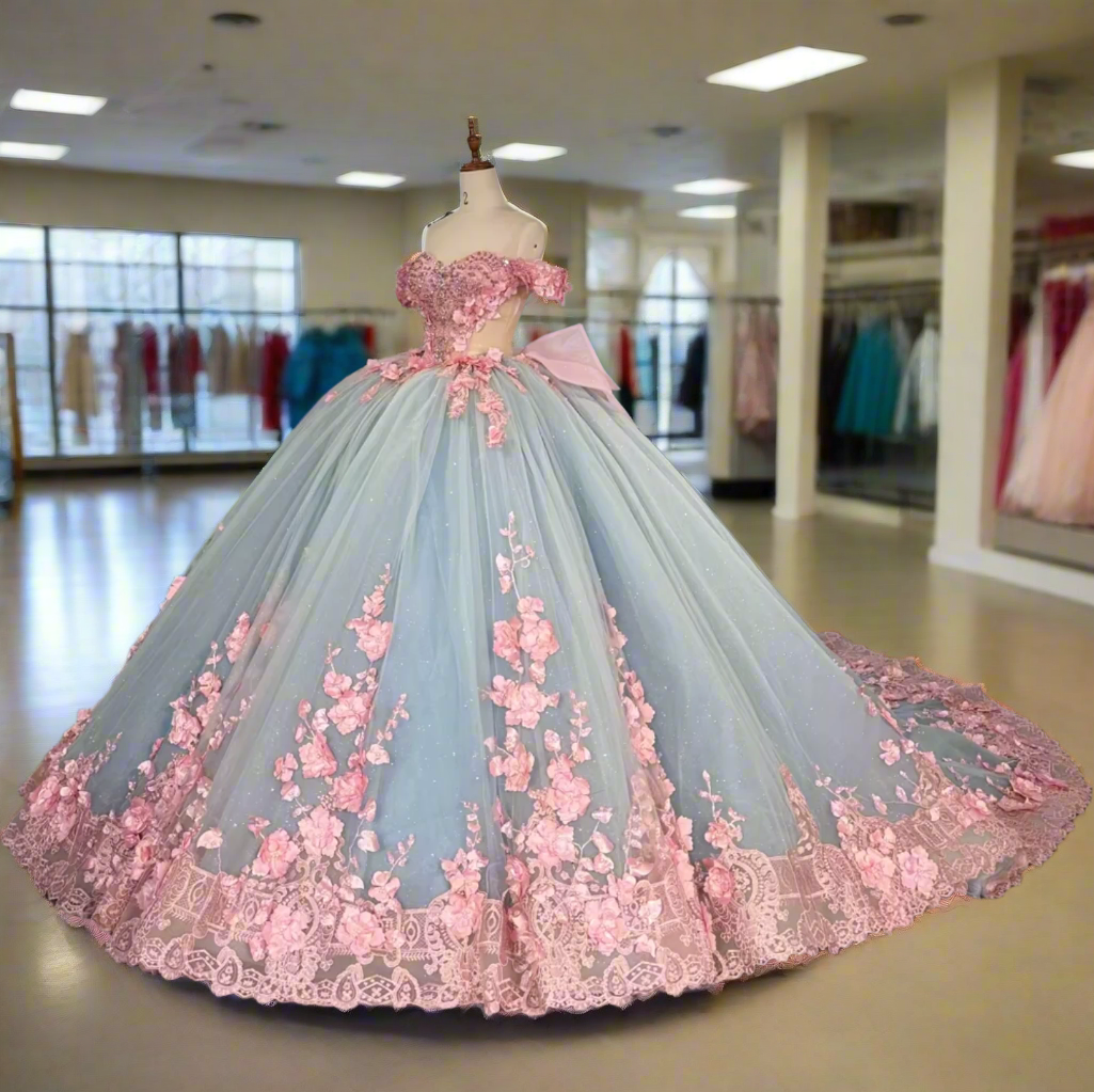 A stunning blue and pink Quinceanera dress with a sheer lace bodice, 3D floral decorations, a sweetheart neckline, and a full ballroom skirt with an oversized back bow.