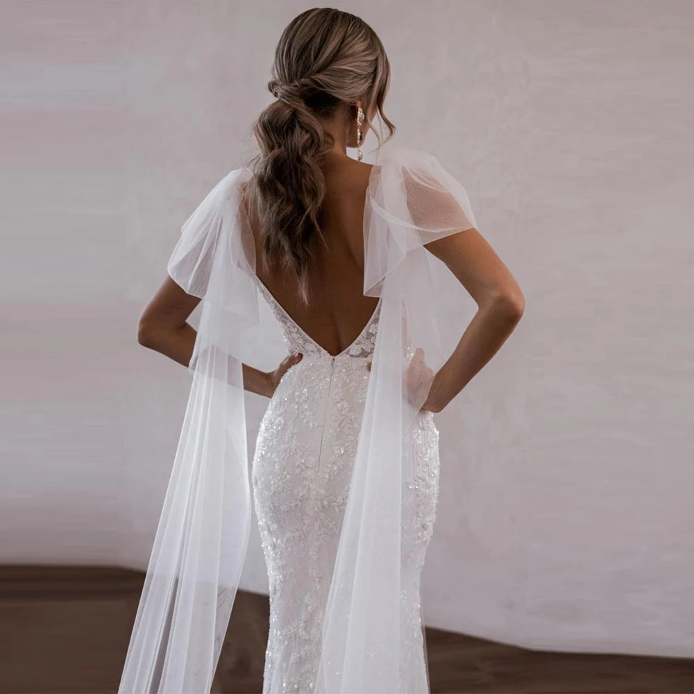 Woman wearing a white wedding dress with a sheer cape against a plain background