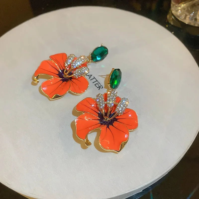 
                      
                        Floral earrings with orange flowers and green gemstones on a white surface.
                      
                    