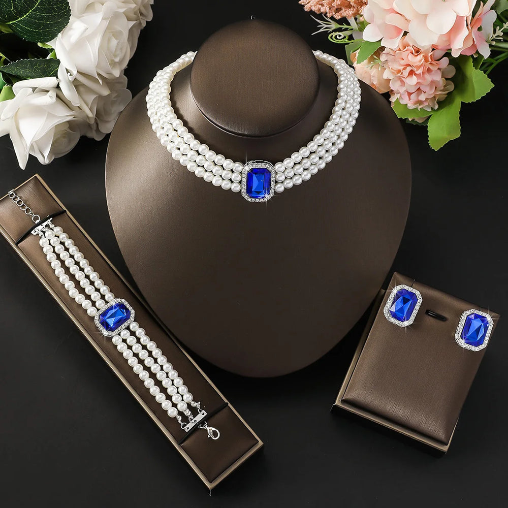 
                      
                        Pearl necklace with blue gemstones on a brown stand, surrounded by flowers.
                      
                    