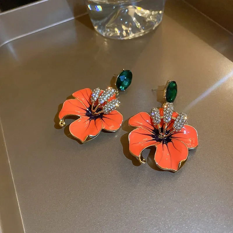 
                      
                        Floral earrings with orange flowers and green leaves on a reflective surface.
                      
                    