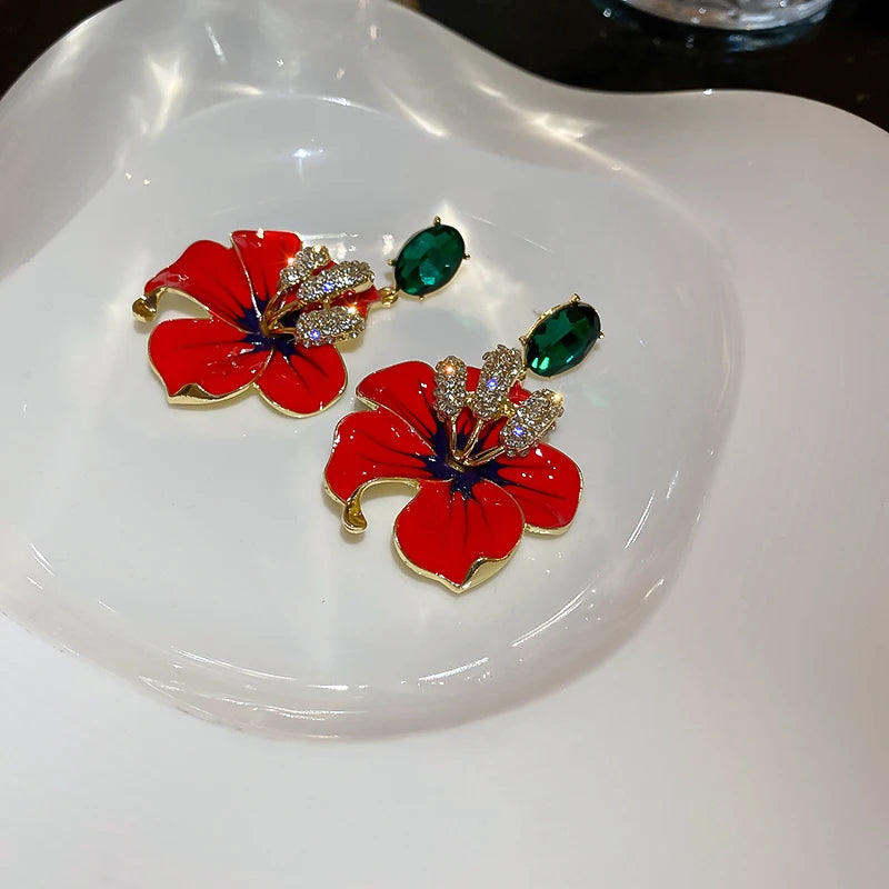 
                      
                        Red flower-shaped earrings with green and gold accents on a white dish.
                      
                    
