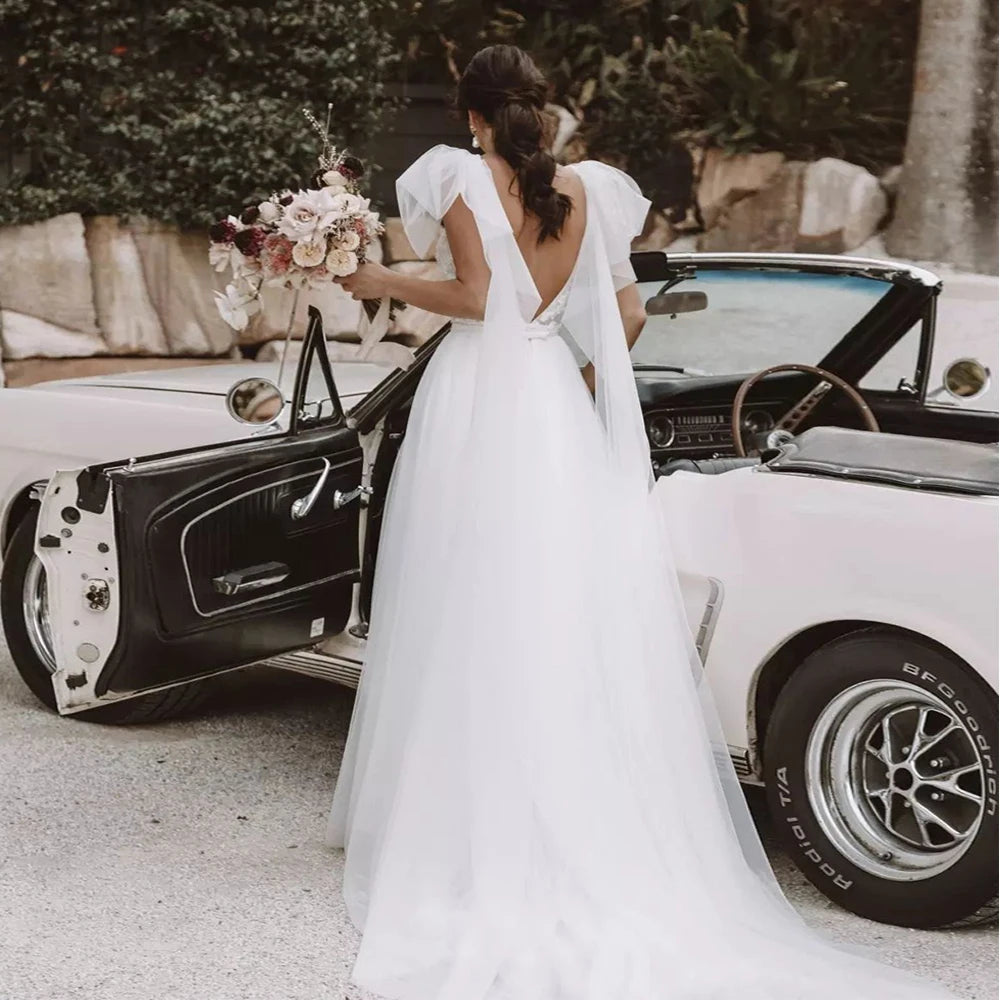 
                      
                        Bride in a white wedding dress standing next to a vintage car with a bouquet of flowers.
                      
                    