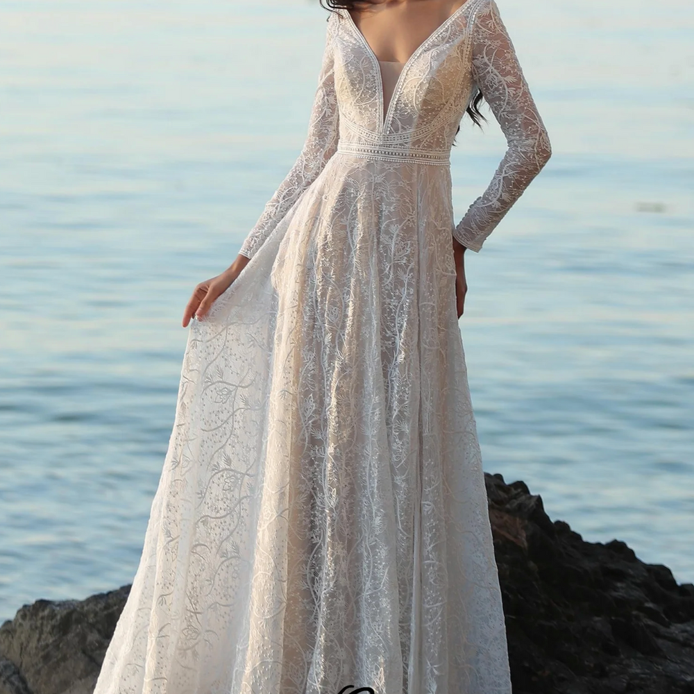 
                      
                        Boho A-Line long sleeve wedding dress with V-neckline, lace details, and illusion back, set against a serene coastal backdrop.
                      
                    