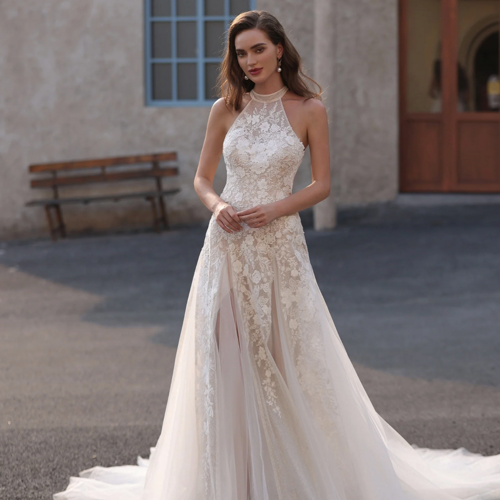 Woman in a white lace wedding dress standing in front of a building with a bench.