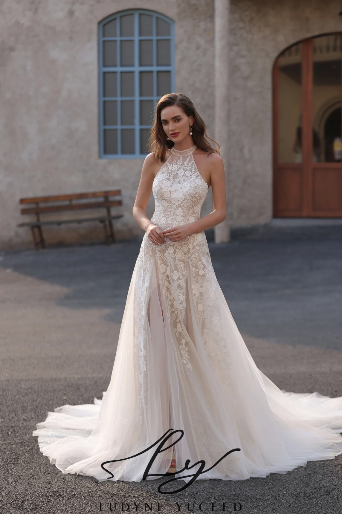 Woman in a white lace wedding dress standing in front of a building with a bench.
