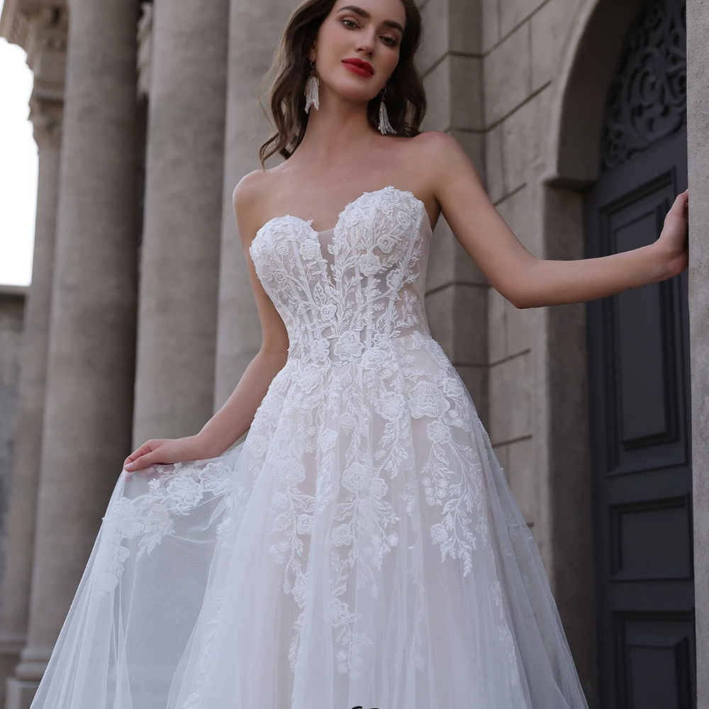 
                      
                        Woman in a white lace wedding dress standing in front of a stone building.
                      
                    
