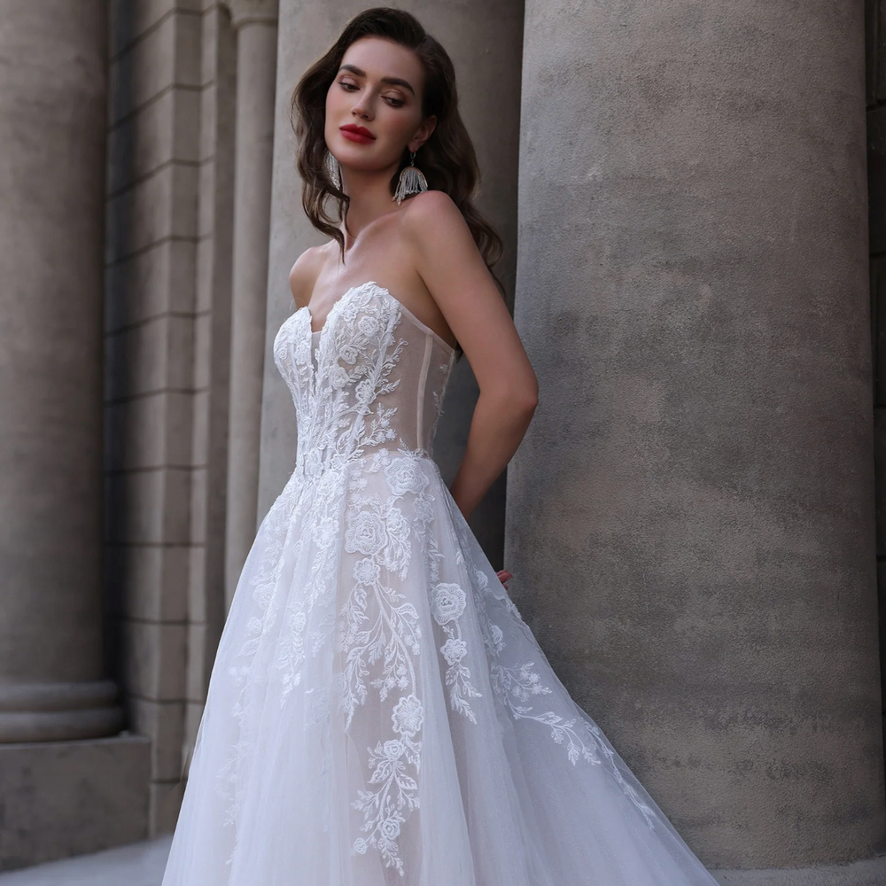 
                      
                        Woman in a white lace wedding dress standing against a stone wall.
                      
                    