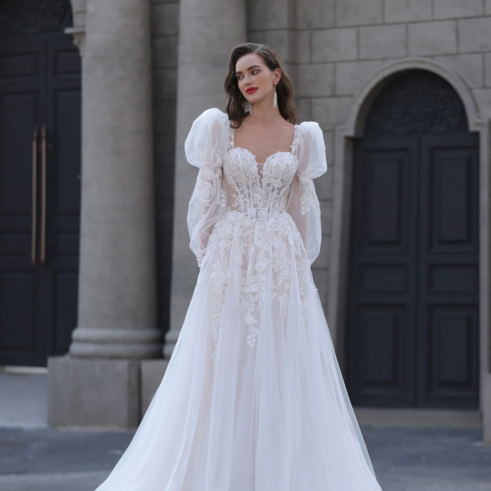 
                      
                        Woman in a white lace wedding dress standing in front of a stone building.
                      
                    