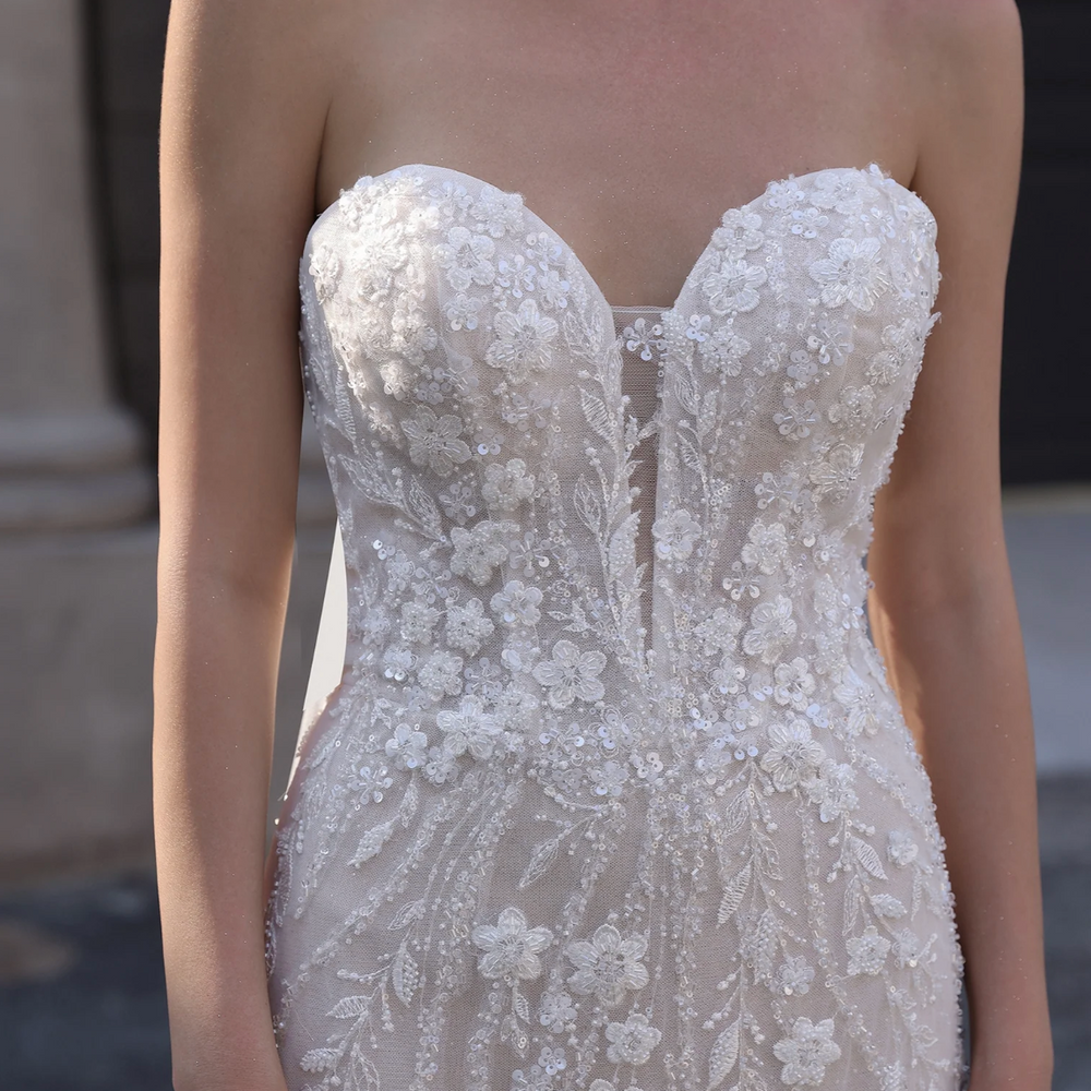 
                      
                        Elegant white strapless gown with floral detailing on a blurred background
                      
                    