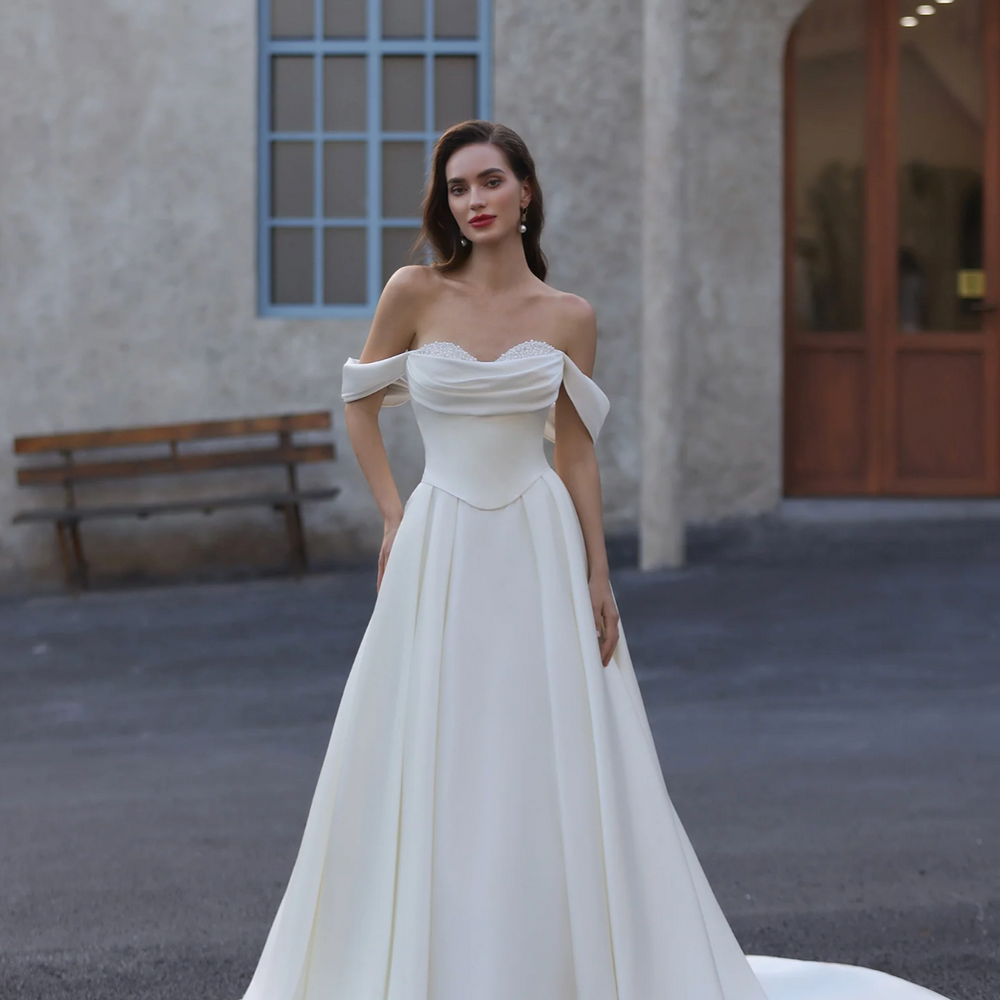 Woman in a white off-shoulder gown standing in front of a building with a bench.