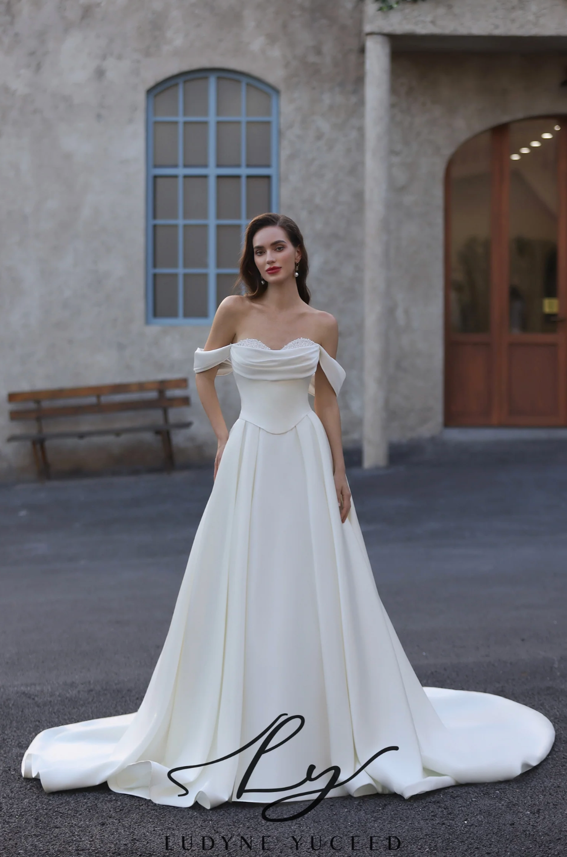 Woman in a white off-shoulder gown standing in front of a building with a bench.