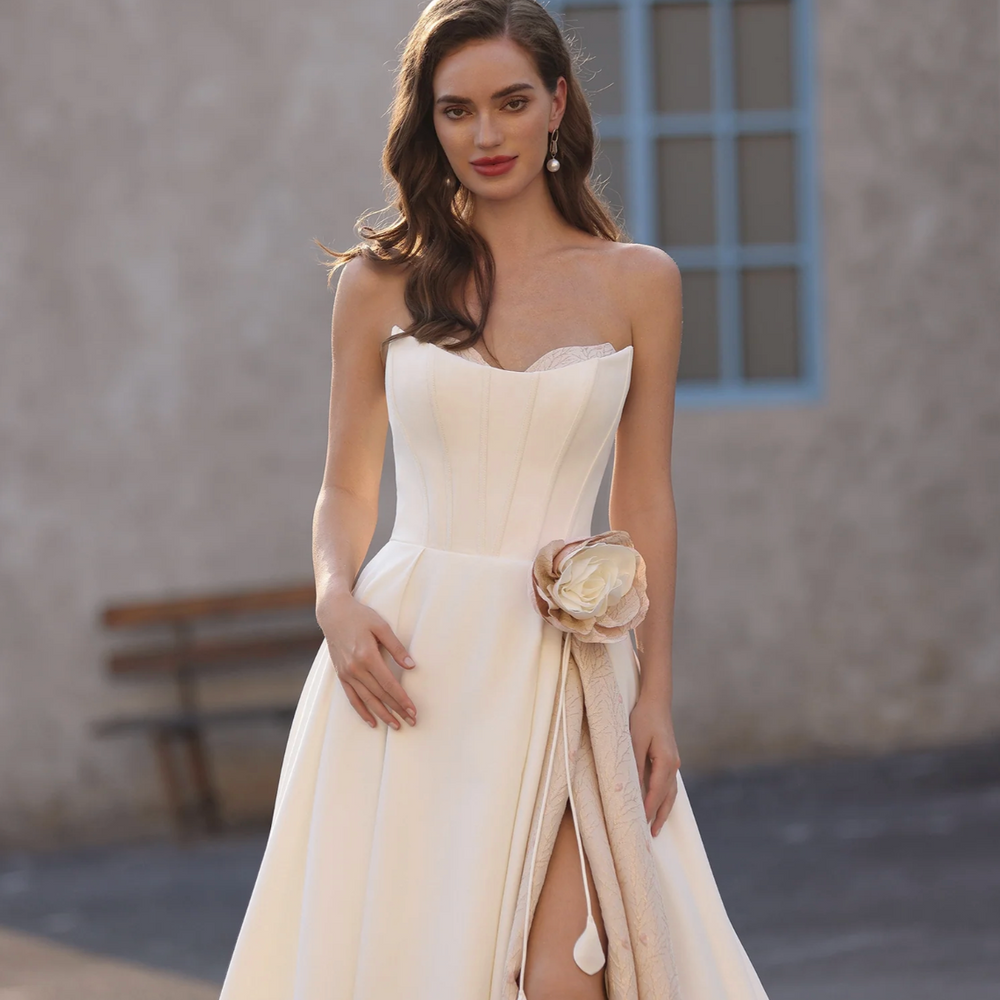 
                      
                        Woman in a white strapless gown with a floral detail, standing against a neutral background.
                      
                    