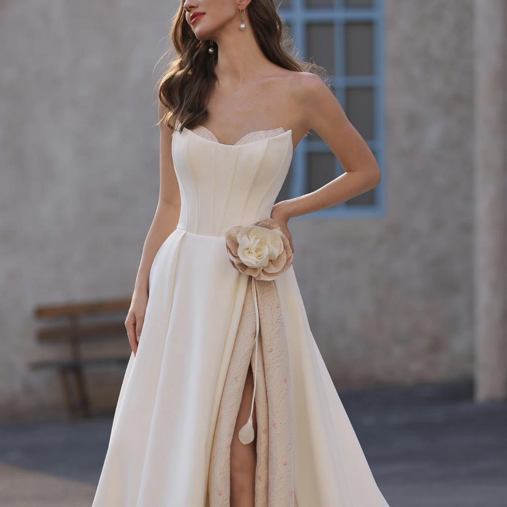 
                      
                        Woman in a white strapless gown with a floral detail, standing outdoors.
                      
                    