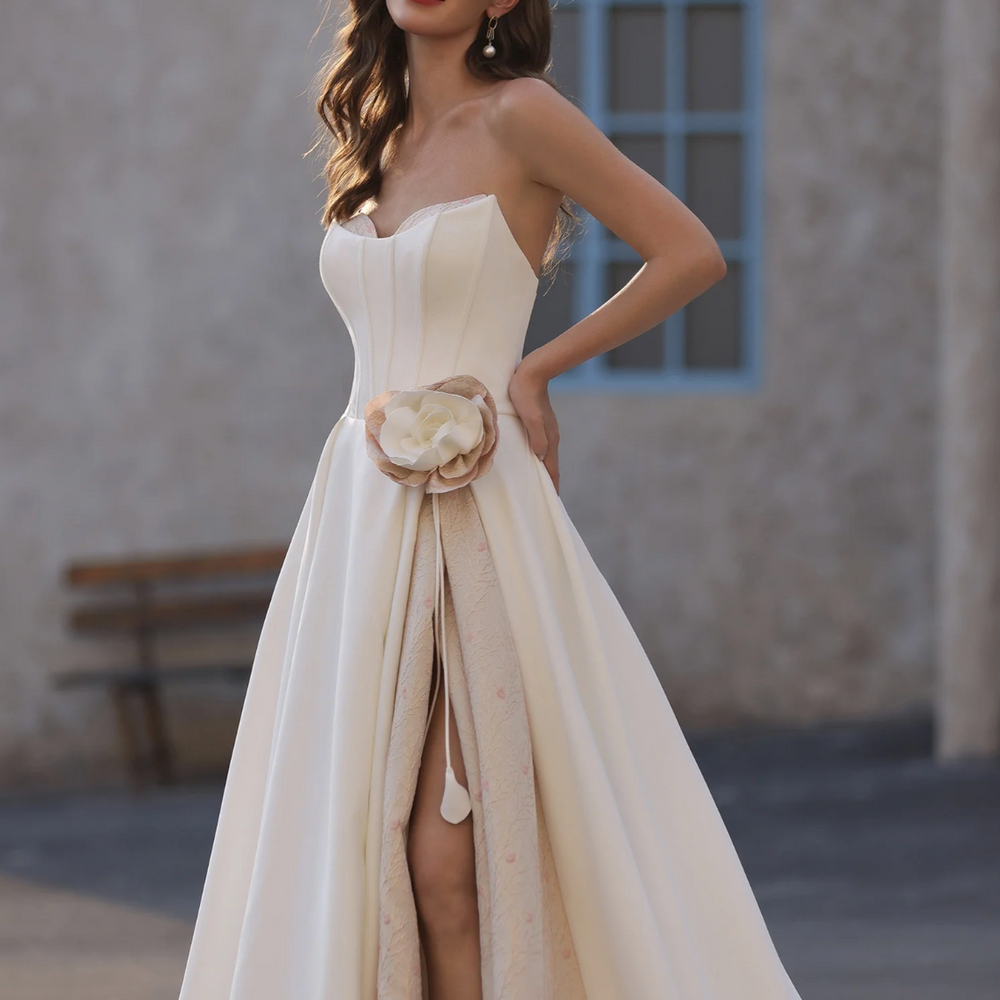 
                      
                        Elegant strapless satin wedding dress with floral accent and thigh-high slit, showcased in a stylish outdoor setting.
                      
                    