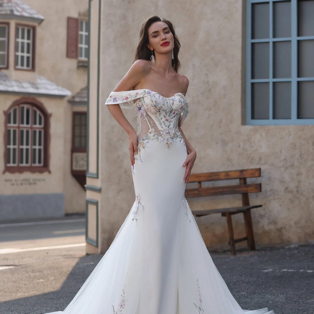 Woman in a white off-shoulder wedding dress standing outdoors with a building in the background.