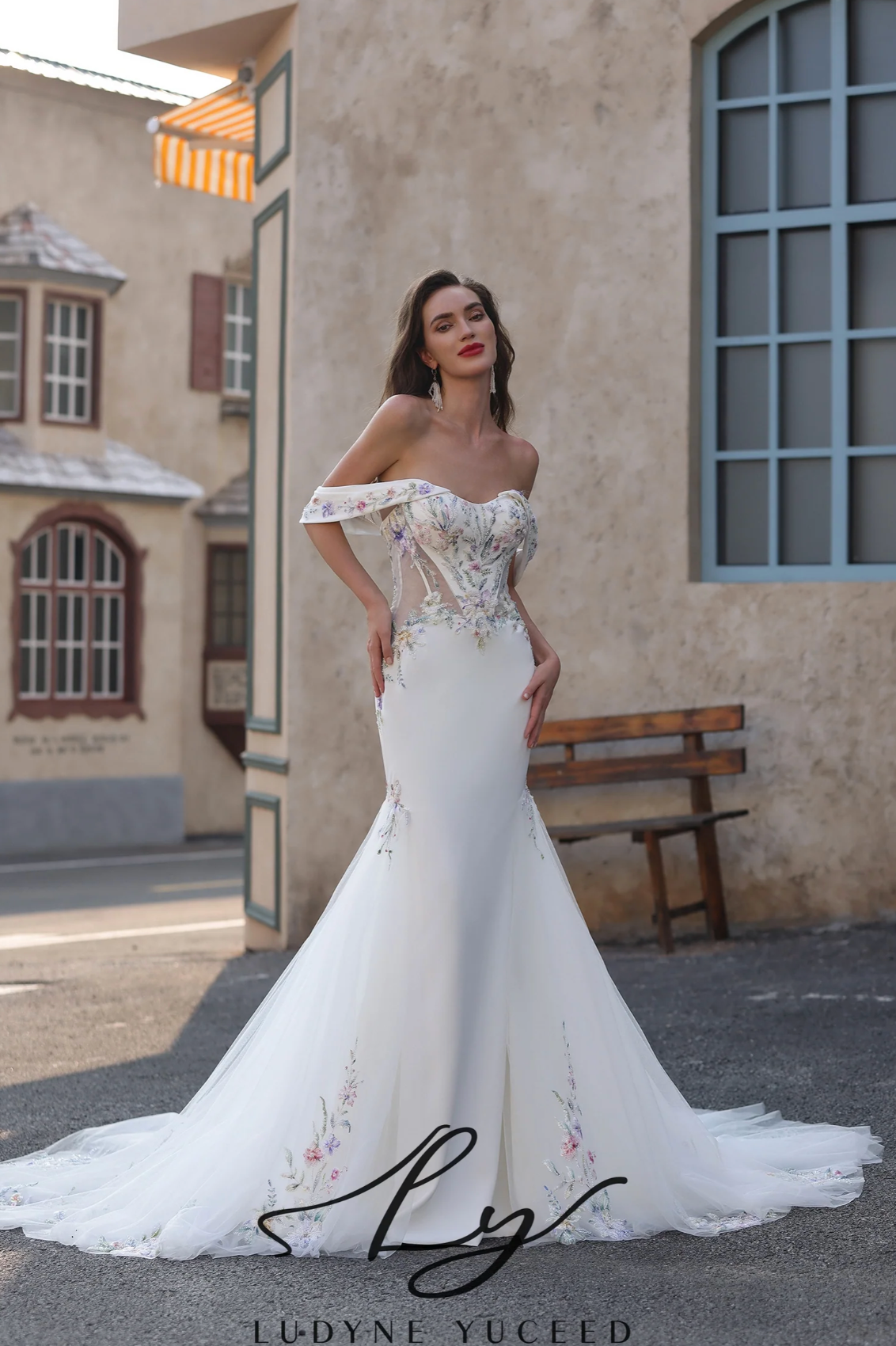 Woman in a white off-shoulder wedding dress standing outdoors with a building in the background.