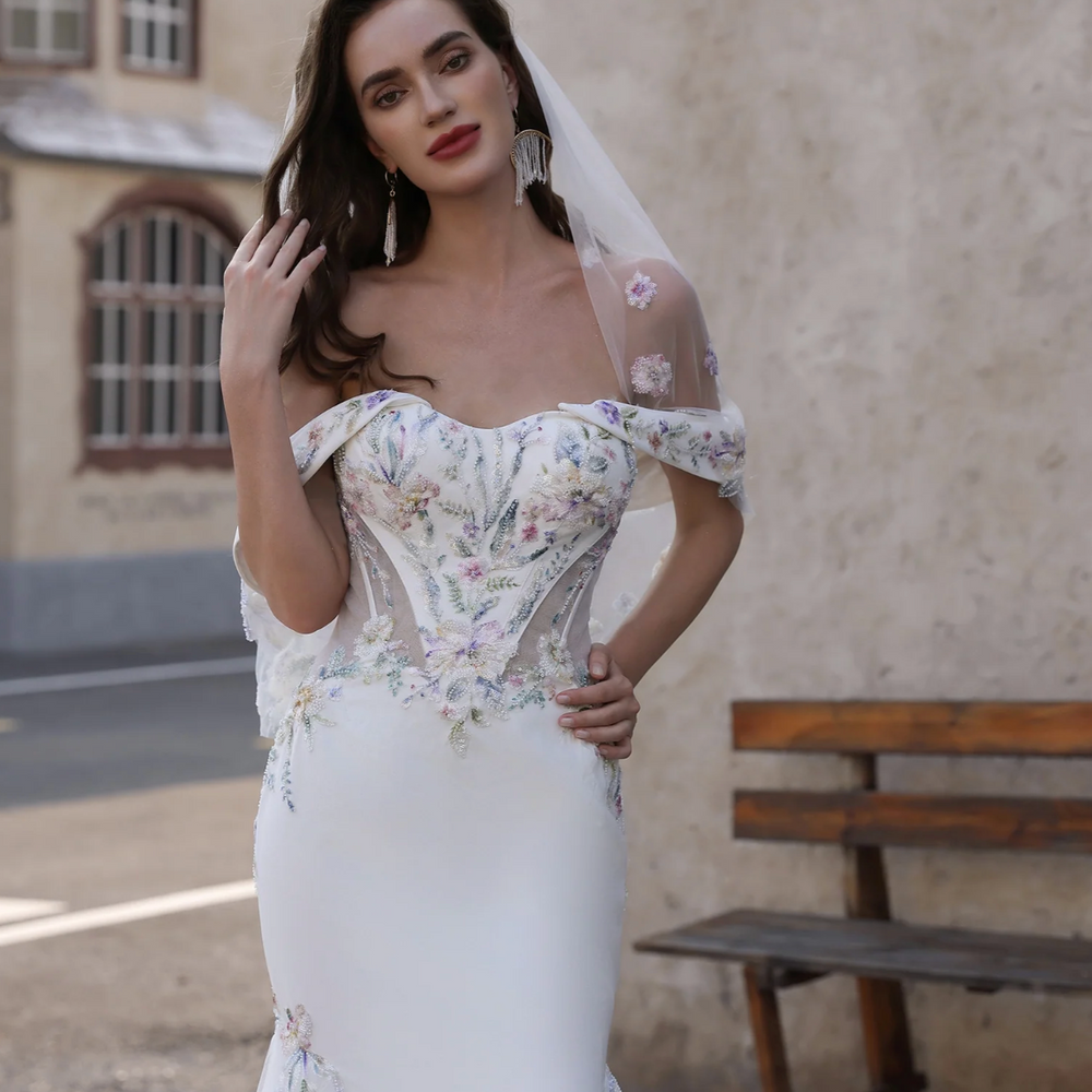 
                      
                        Woman in a floral off-shoulder wedding dress with a veil, standing outdoors.
                      
                    