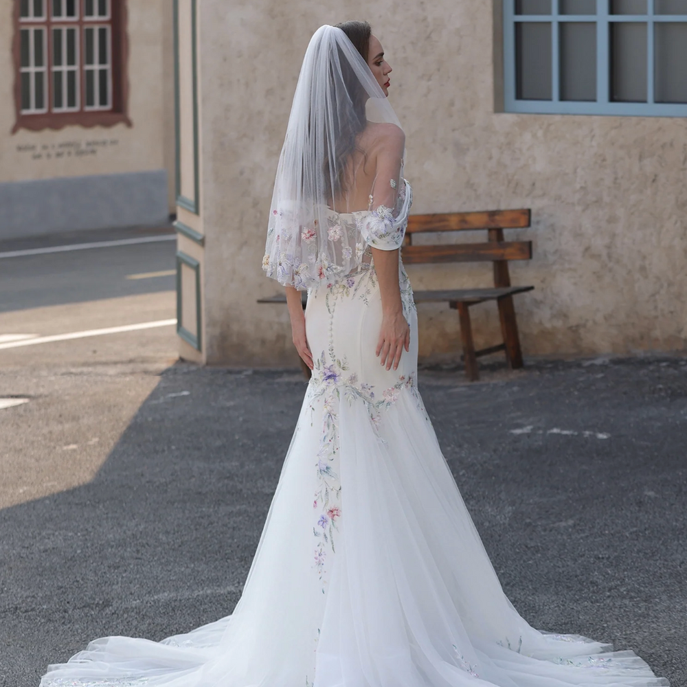 
                      
                        Woman in a white wedding dress with a floral veil standing outdoors near a building.
                      
                    