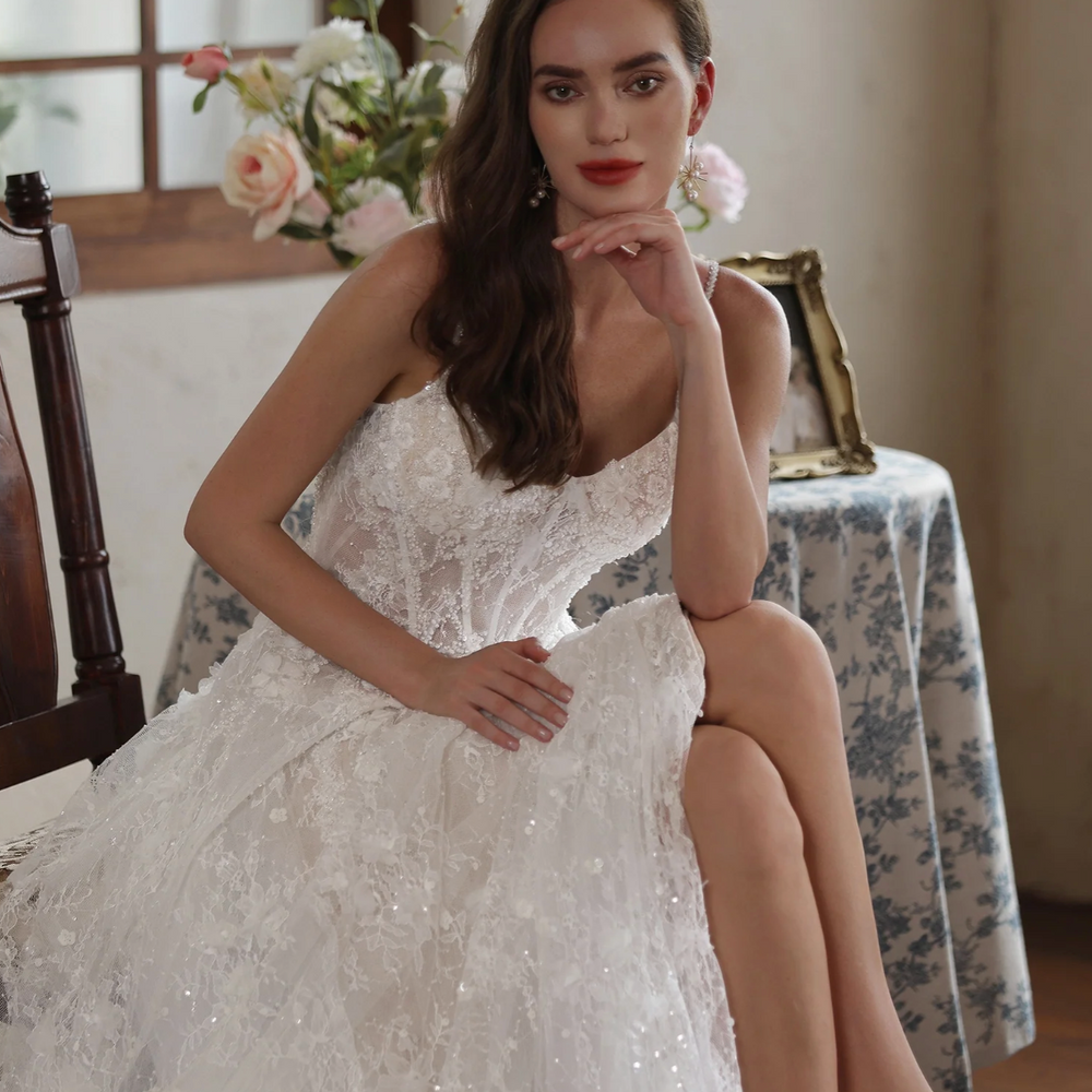 
                      
                        Elegant model wearing a romantic floral lace wedding gown with delicate straps, seated in a charming setting.
                      
                    
