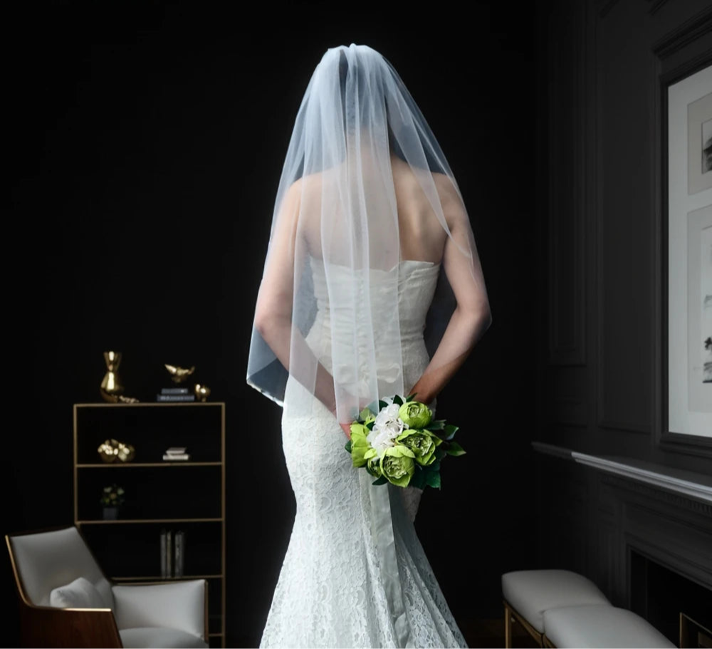 
                      
                        Woman in a wedding dress with a veil holding a bouquet in a room with dark walls and minimal decor.
                      
                    